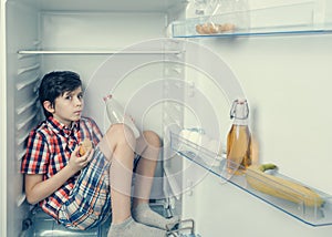 An alert boy in a shirt and shorts eating a croissant and drink milk inside a fridge with food and product. Close-up