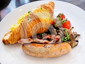 All-Day breakfast / brunch meal platter of scrambled eggs; croissant; sausage; bacon and mushrooms on a white plate