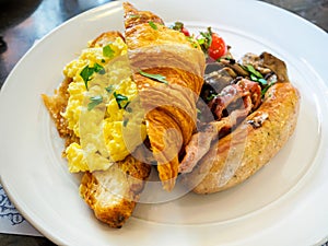 All-Day breakfast / brunch meal platter of scrambled eggs; croissant; sausage; bacon and mushrooms on a white plate