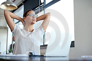 All in a days work. a young woman leaning back in her chair with her hands behind her head working on a laptop in an