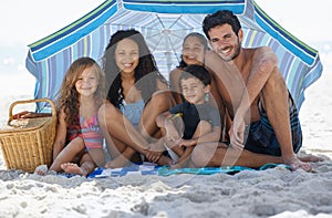 They all love the beach. A happy family smiling at the camera while sitting under an umbrella at the beach.