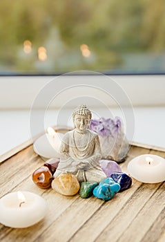 All seven chakra colors crystals stones around sitting Buddha figurine on natural wooden tray.