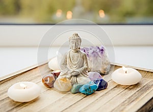 All seven chakra colors crystals stones around sitting Buddha figurine on natural wooden tray.