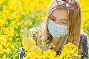 Allergic blonde girl in a protective mask looks at the yellow flower sitting on a meadow