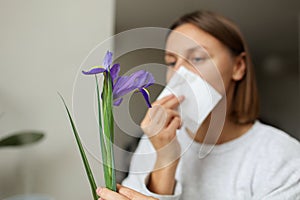 Allergic young woman holds iris flower, covers nose with paper tissue has runny nose, sneezes from flowers pollen at