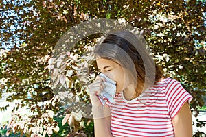 Allergy concept. Sneezing young girl with nose wiper among blooming trees in park