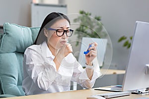 Allergy, feeling unwell at the workplace. A young Asian woman uses an inhaler in the office