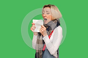 Allergy, runny nose. Portrait of sick adult woman wearing warm scarf standing with opened mouth to sneeze in napkin, feeling