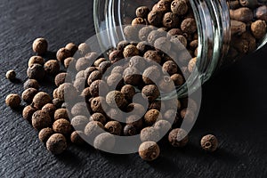 Allspice jamaica pepper grains spilled out of the glass jar over black slate background. Dried berries of the Pimenta dioica tree