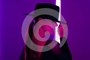 Alluring woman in hat between flashing colored neon lights, studio background. Femme fatale, stylish outfit of ballet