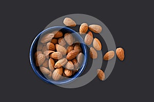 Almonds lying in a blue bowl isolated on dark background with almonds scattered around. Top view image