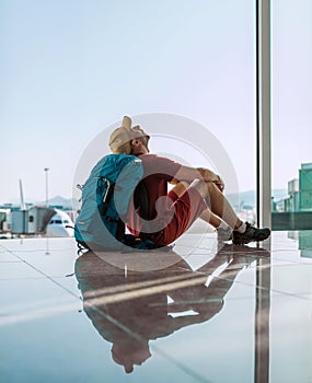 Alone backpacker traveler sitting on the airport terminal floor near the window, looking at the hall ceiling and waiting for