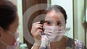 Alone Caucasian young woman in protective medical pink mask in night robe makeup drawing paint brush her eyebrow