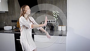 Alone crazy brunette woman is wearing pajamas and dancing in kitchen in evening, shaking her hands and body