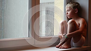 Alone little boy looks raindrops through window glass