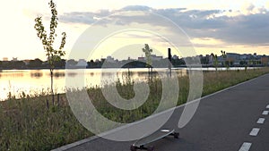 Alone longboard on asphalt road path on river bank in sunset in park out of city with skyline