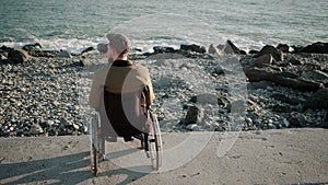 Alone male wheelchair user is sitting calmly on seacoast and relaxing, back view