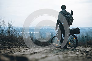 Alone man with a backpack and a bicycle stands and looks into the distance. hilltop overlooking a valley in haze, a city on the