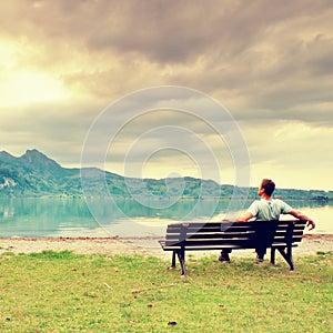 Alone man sits on bench beside an azure mountain lake. Man relax