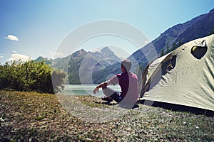 Alone man travel near the lake mountain background, travel alone concept. Young tourist guy sitting after a green tent and looking