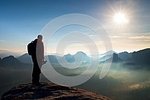 Alone photographer with heavy backpack and tripod in hand on rocky cliff and watching down to deep misty valley bellow