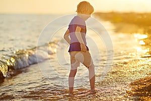 Alone preschooler boy walking barefoot along the seashore during summer holidays. Child having fun in waves on sunset. Family