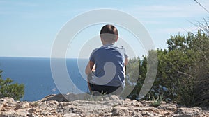 Alone sad boy sitting on the ground and throws stones into water at sunny day