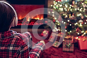 Alone sad woman in headphones sitting and warming at winter evening near fireplace flame and  christmas tree.