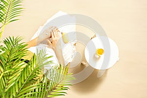 Alone woman applying sunscreen for protection skin sitting under palm tree branches near beach table with orange juice. Top view.