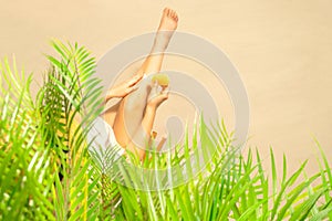 Alone woman applying sunscreen on skin sitting under palm tree branches. Female relaxation on the sand of the beach at summer