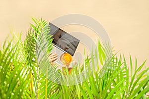 Alone woman freelancer holding glass of water with piece orange working on laptop sitting under palm tree branches on the sand