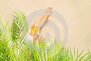 Alone woman sitting under palm tree branches with glass of water with piece orange. Female relaxation on the sand of the beach