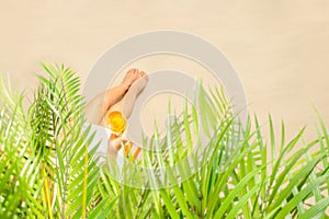 Alone woman sitting under palm tree branches with glass of water with piece orange. Female relaxation on the sand of the beach