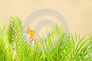 Alone woman sitting under palm tree branches with glass of water with piece orange. Female relaxation on the sand of the beach