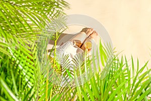 Alone woman sitting under palm tree branches with glass of water with piece orange. Female relaxation on the sand of the beach