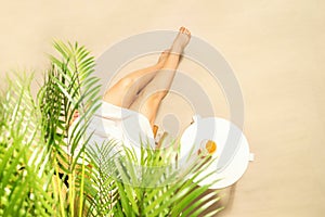 Alone woman sitting under palm tree branches. Glass of water with piece orange on the table. Female relaxation on the sandy beach.