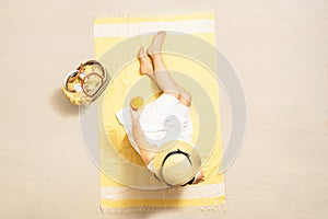 Alone woman in straw hat and dress sitting on yellow beach towel with bag, headphones, and orange juice. Summer vacation.