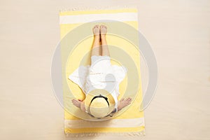 Alone woman in straw hat and dress sitting on yellow beach towel. Female relaxation on the sand of the beach at summer vacation.