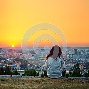 Sama mladá dívka sedící nad městem a při pohledu na klidný letní západ slunce