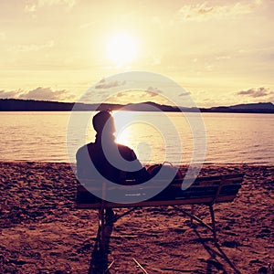 Alone Young Man In Silhouette Sitting In The Sun On Beach. Tourist take rest on wooden bench at autumn lake.