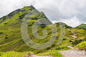 Alpine hiking trail. A stone winding road, surrounded by green plants, alpine roses. The cross at the top of the mountain