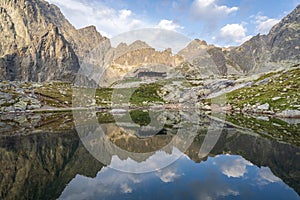 Vysokohorská chata obklopená horami odrážajúcimi sa v popredí plesa pri východe slnka, Slovensko, Európa