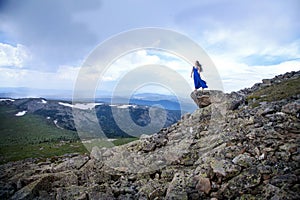 An Altai girl in a blue dress stands on top of the Sarlyk mountain above a cliff among rocks and stones. Tourism concept