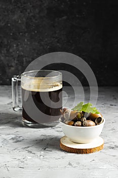 Alternative non-caffeinated drink in a glass mug with ripe acorns in a white plate on a dark background