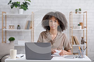 Alternative work at home. Cute african american woman with notepad looks at laptop