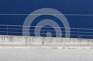 An aluminum blue cladding wall with an oblique railing in front of a sidewalk and an asphalt street.