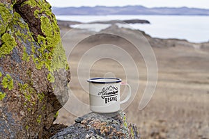 Aluminum enameled mug of a traveler with hot tea on a moss-covered mountain on a picturesque background of Lake Baikal.