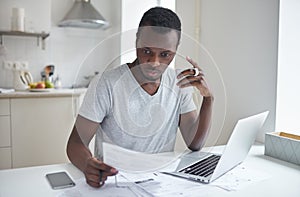 Amazed african man sits at kitchen table, holds cup of coffee, looks at bills