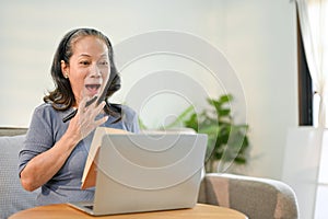 Amazed Asian-aged retired woman covering her mouth, looking at her laptop screen