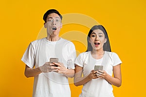 Amazed asian couple holding smartphones over yellow background